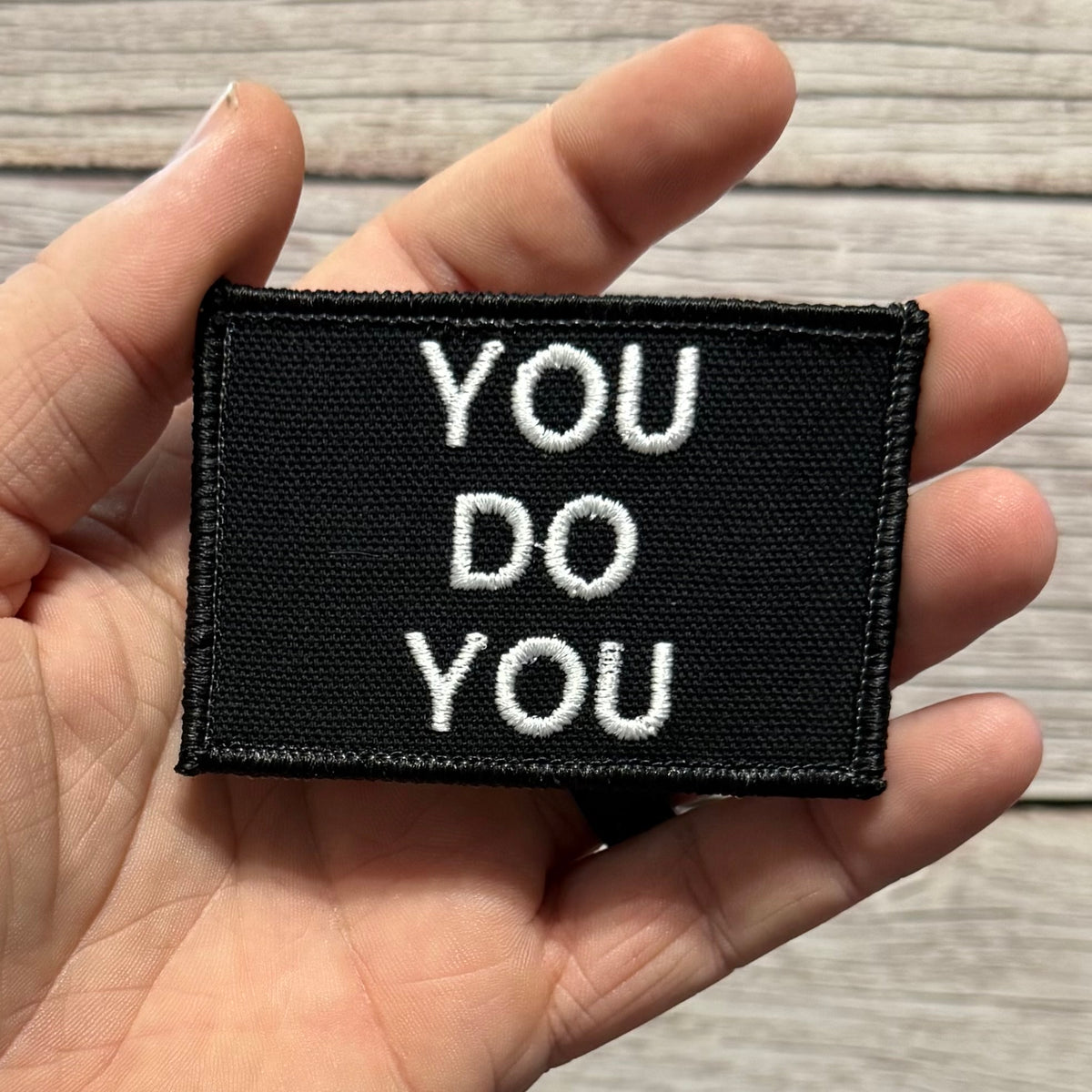 A black rectangular embroidered patch with the words 'YOU DO YOU' in white capital letters, held in a person's hand against a light wood background.