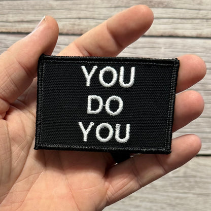 A black rectangular embroidered patch with the words 'YOU DO YOU' in white capital letters, held in a person's hand against a light wood background.