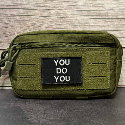 An olive green tactical pouch with a black rectangular patch that reads 'YOU DO YOU' in white capital letters, set against a wood panel background.
