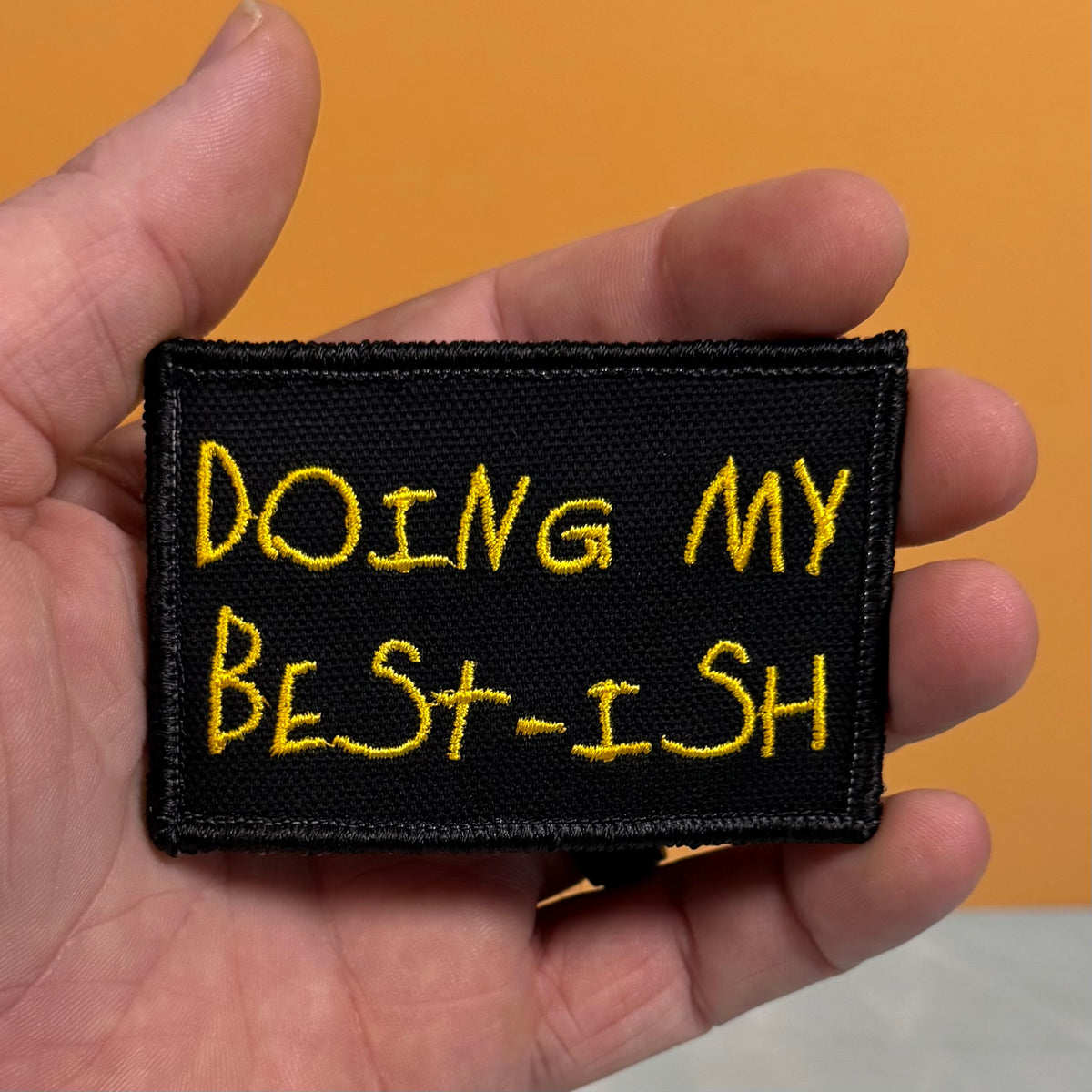 A hand holding a rectangular black embroidered patch with yellow text that reads 'DOING MY BEST-ISH' against a solid orange background.