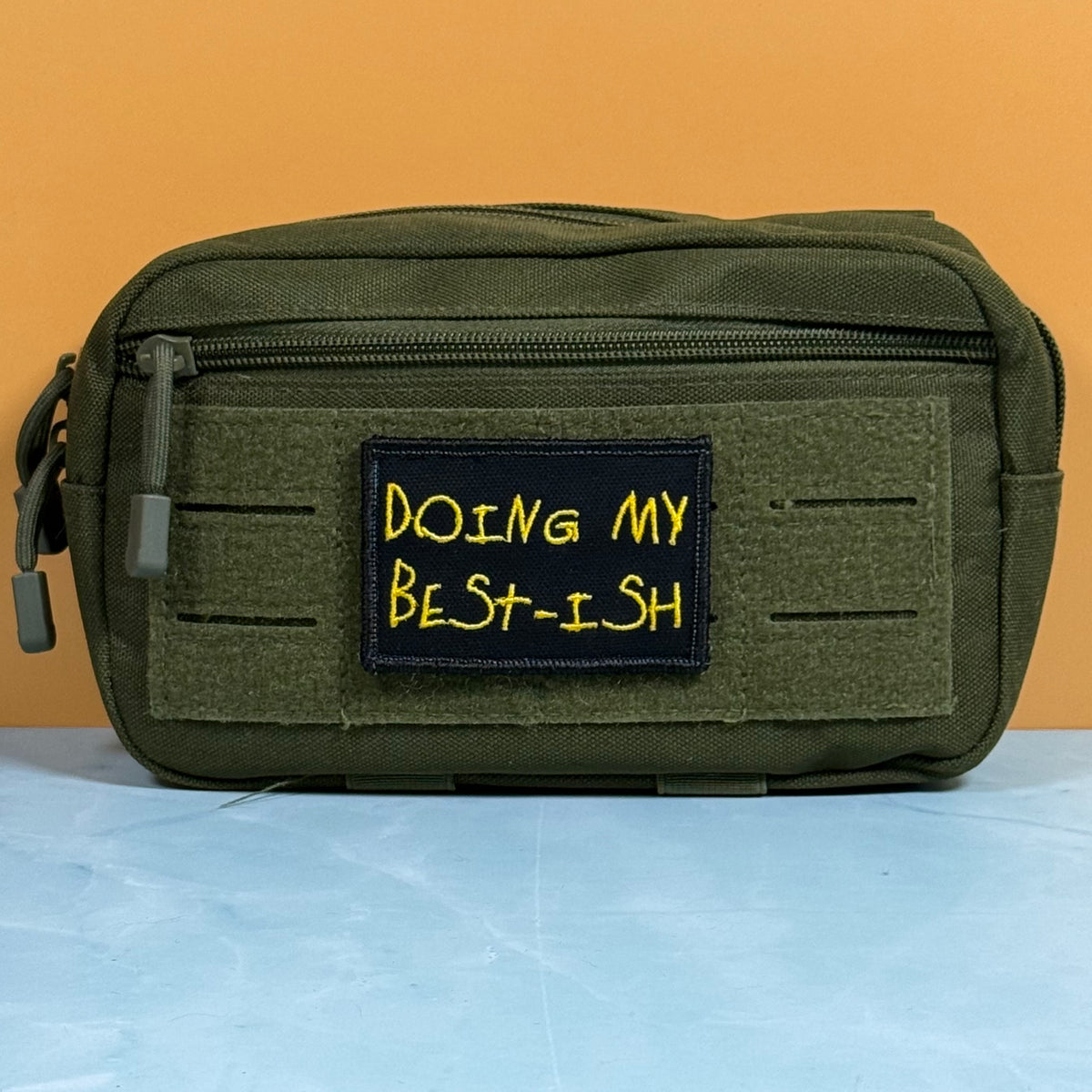 An olive green tactical pouch with a front zipper and a black patch embroidered with yellow text reading 'DOING MY BEST-ISH', set against a solid orange background.