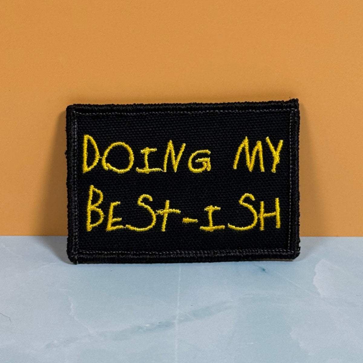 A rectangular black embroidered patch with yellow text reading 'DOING MY BEST-ISH' against a two-tone orange and light gray background.