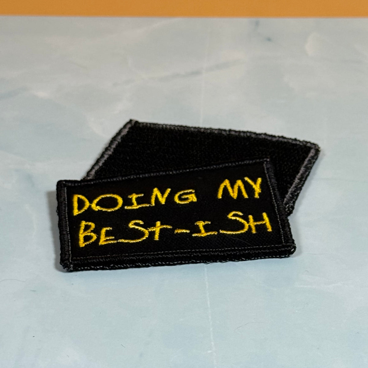 Two black embroidered patches with yellow text, one reading 'DOING MY BEST-ISH', displayed on a light gray marble-like surface.