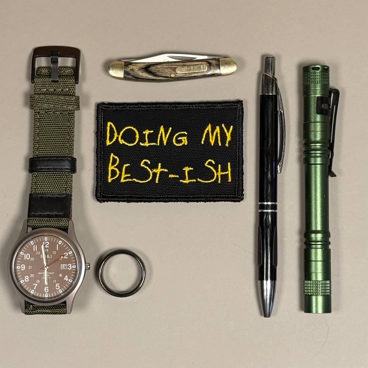 A flat-lay arrangement of various tactical and utility accessories on a neutral beige background, including an olive green watch with a black dial, a black embroidered patch with yellow text reading 'DOING MY BEST-ISH', a black pen, a green tactical flashlight, a pocket knife, and a black metal ring.