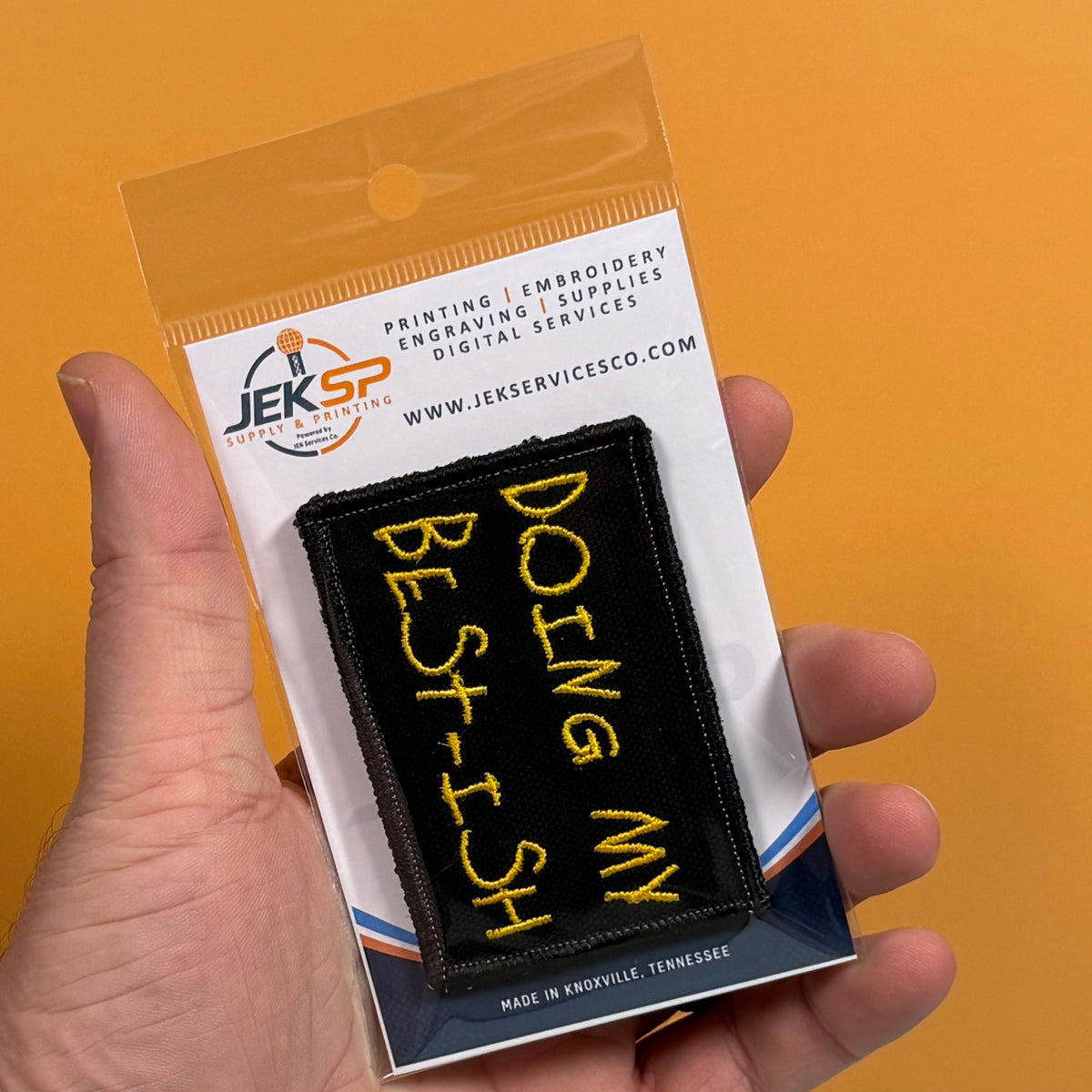 A hand holds a small black embroidered patch with yellow text reading 'DOING MY BEST, ISH' inside its clear plastic retail packaging against a solid orange background.