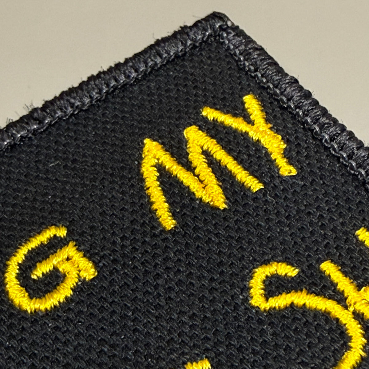 Close-up of a black embroidered patch with yellow lettering on a light beige background.