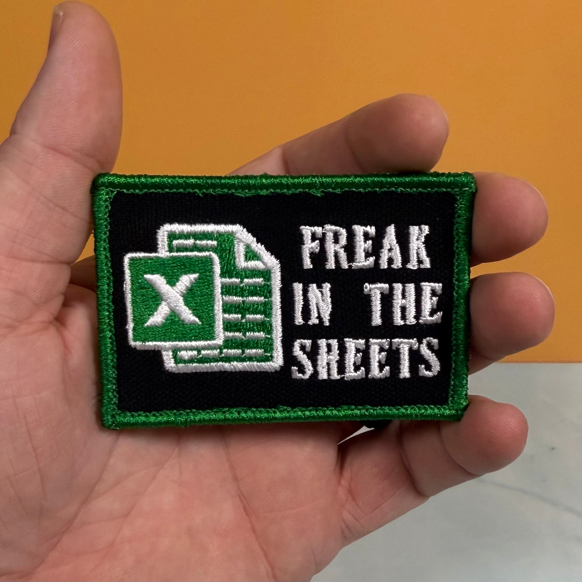 A hand holding a rectangular embroidered patch with a black background, green border, a green Excel-style icon, and the white text 'FREAK IN THE SHEETS' against an orange background.