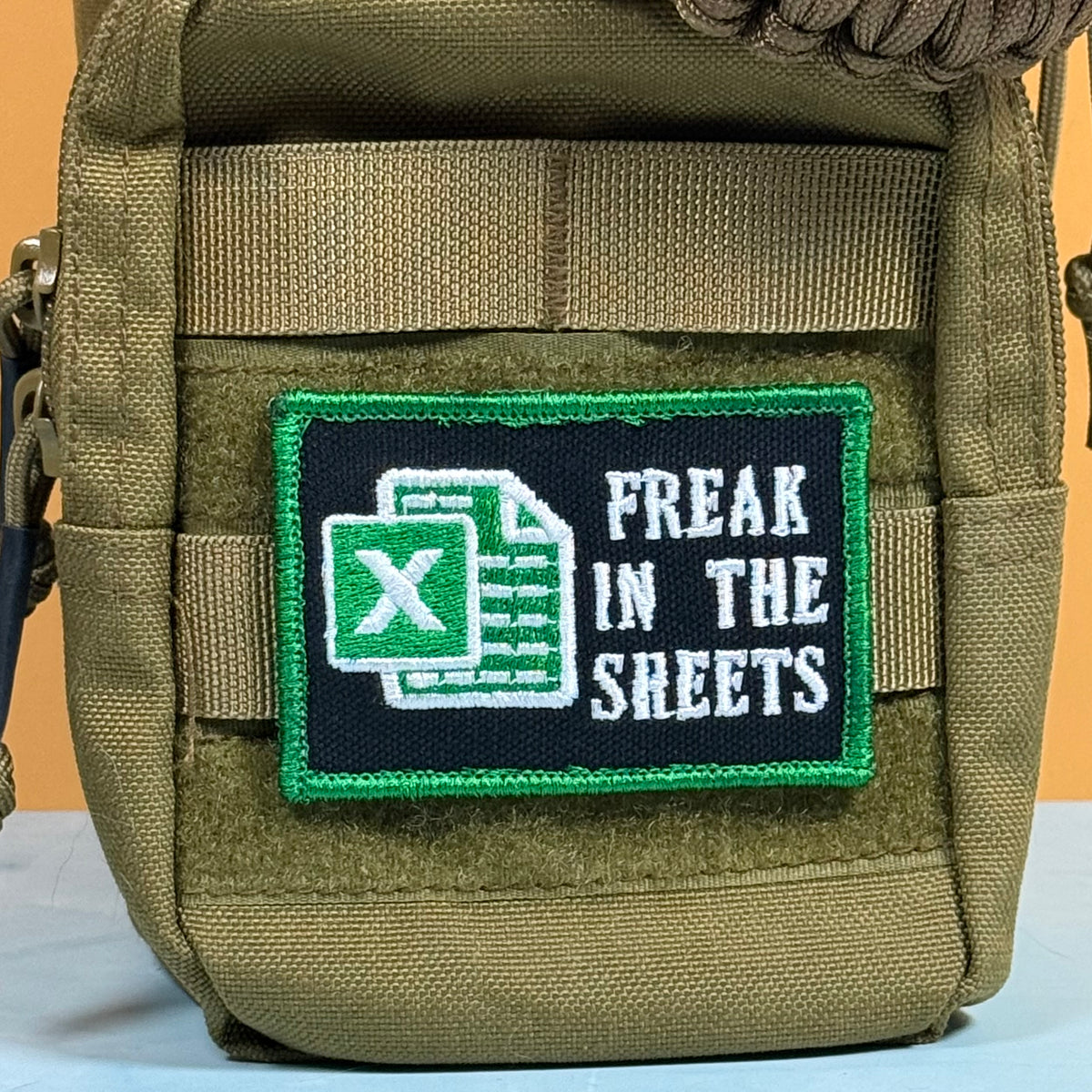 An olive drab tactical pouch featuring a rectangular embroidered patch with a black background, green border, white Excel icon, and the text 'FREAK IN THE SLEETS'.