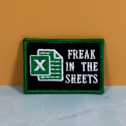 An embroidered patch with a green border, black background, a green Excel-style sheet icon, and the words 'FREAK IN THE SHEETS' in white, displayed against an orange wall and light gray surface.