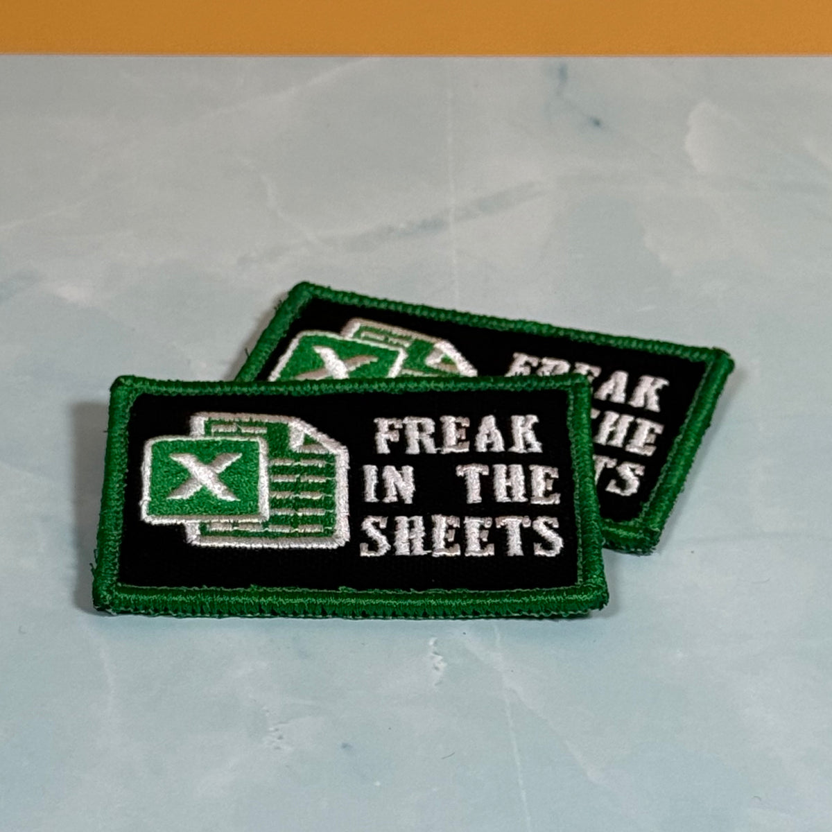 Two embroidered rectangular patches with a black background, green border, and a green Excel-style spreadsheet icon next to the white text 'FREAK IN THE SHEETS', displayed on a light marble surface.