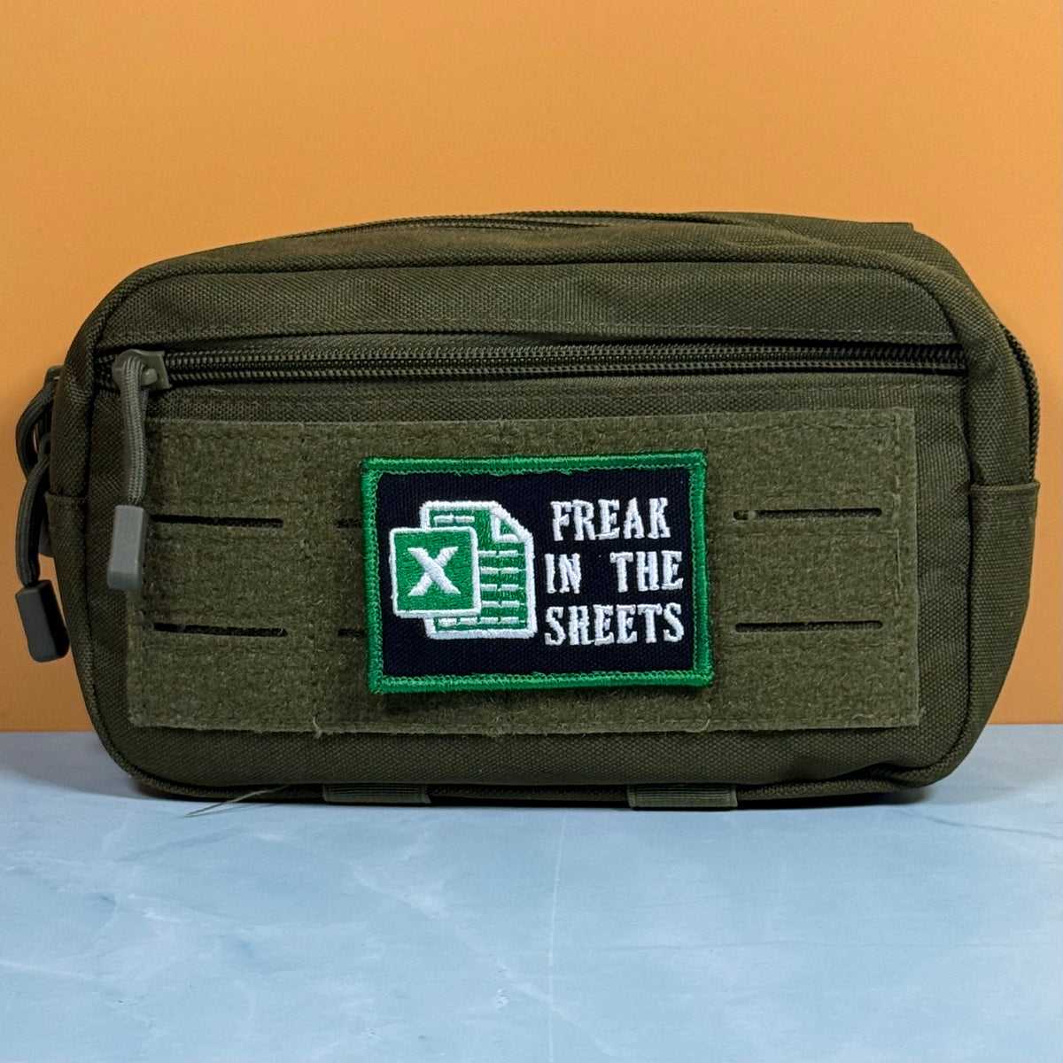 An olive green tactical pouch with a front hook-and-loop patch that reads 'FREAK IN THE SHEETS' alongside a green Excel logo, set against a solid orange background.