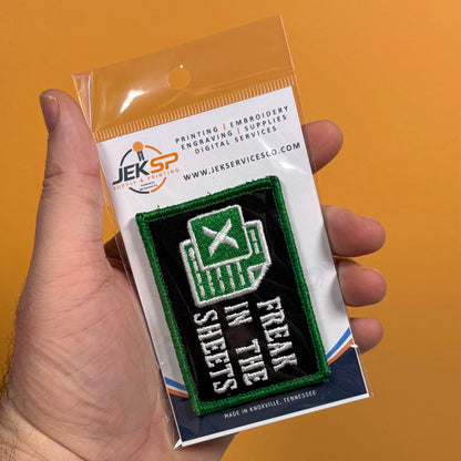 A hand holds a small embroidered patch on a clear plastic backing card against a solid orange background. The patch is black with green border and white text reading 'FREAK IN THE SHEETS,' with a green Excel-style spreadsheet icon.