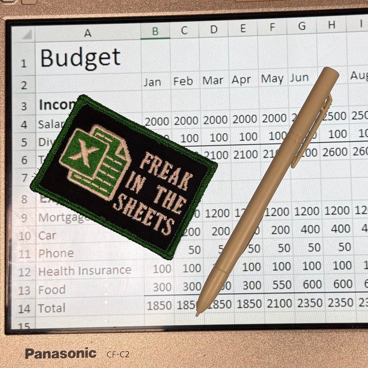 A rectangular embroidered patch with a black background, green border, and a green spreadsheet icon, reading 'FREAK IN THE SHEETS,' is placed on top of a budget spreadsheet displayed on a Panasonic CF-C2 tablet. A beige pen rests on the right side.