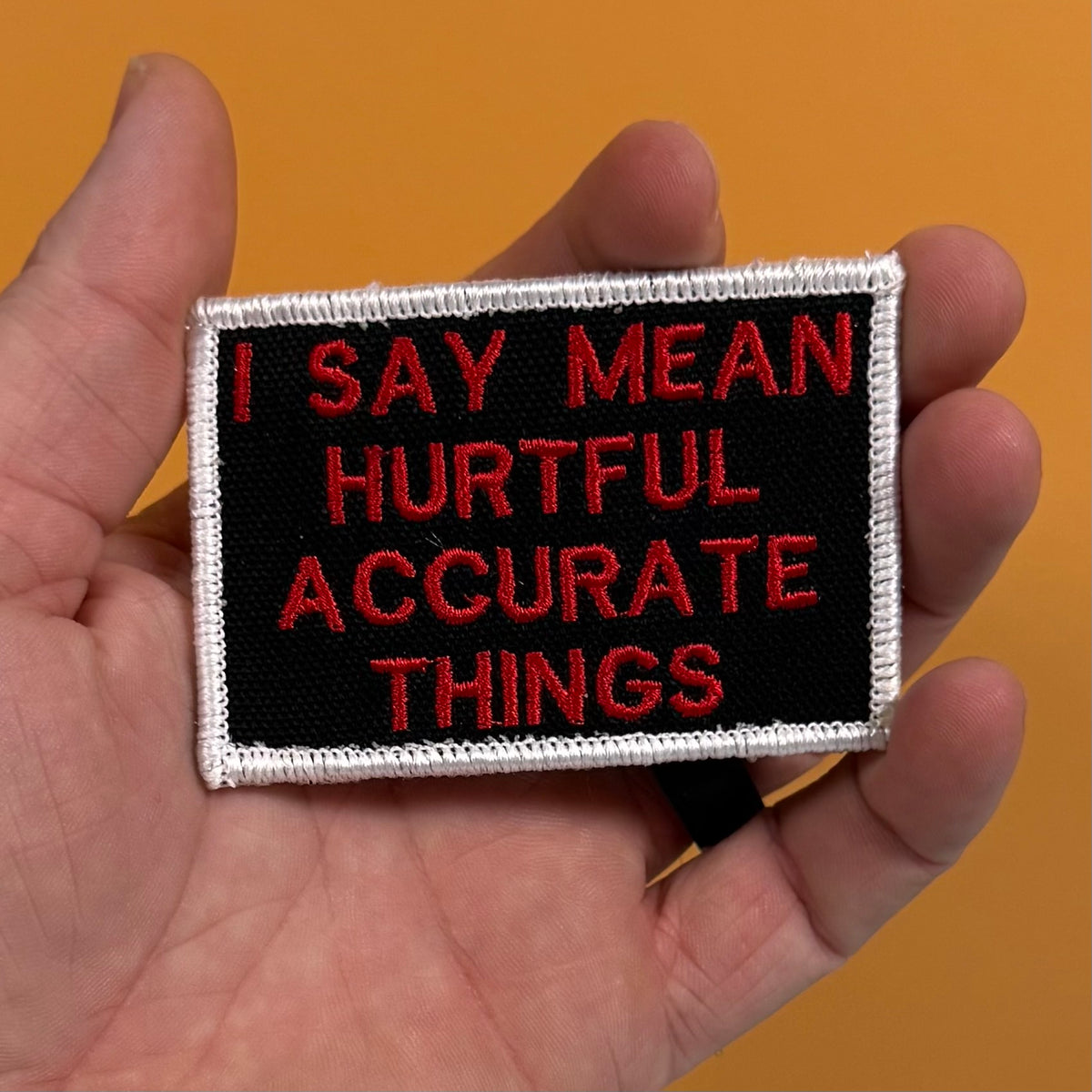 A hand holding a rectangular embroidered patch with a black background, white border, and red text that reads, 'I SAY MEAN HURTFUL ACCURATE THINGS' against a solid orange background.