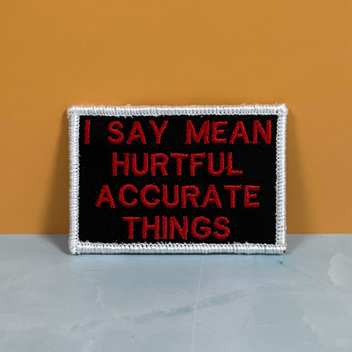 A rectangular embroidered patch with a black center, red text reading 'I SAY MEAN HURTFUL ACCURATE THINGS,' and a white stitched border, photographed against a mustard-yellow wall and pale gray surface.