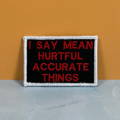 A rectangular embroidered patch with a black center, red text reading 'I SAY MEAN HURTFUL ACCURATE THINGS,' and a white stitched border, photographed against a mustard-yellow wall and pale gray surface.