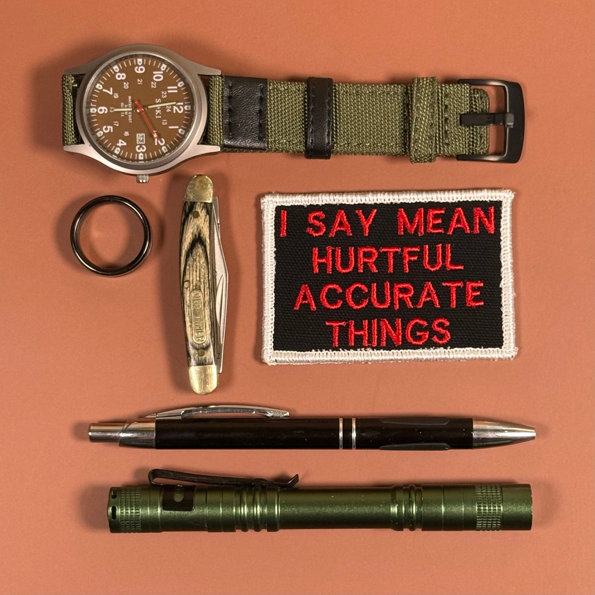 A flat-lay collection of outdoor and survival gear on a solid terracotta background, including an olive green tactical watch, a folding pocket knife, a black ring, a black and red embroidered patch reading 'I SAY MEAN HURTFUL ACCURATE THINGS', a black and silver pen, and an olive green flashlight.