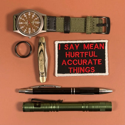 A flat-lay collection of outdoor and survival gear on a solid terracotta background, including an olive green tactical watch, a folding pocket knife, a black ring, a black and red embroidered patch reading 'I SAY MEAN HURTFUL ACCURATE THINGS', a black and silver pen, and an olive green flashlight.