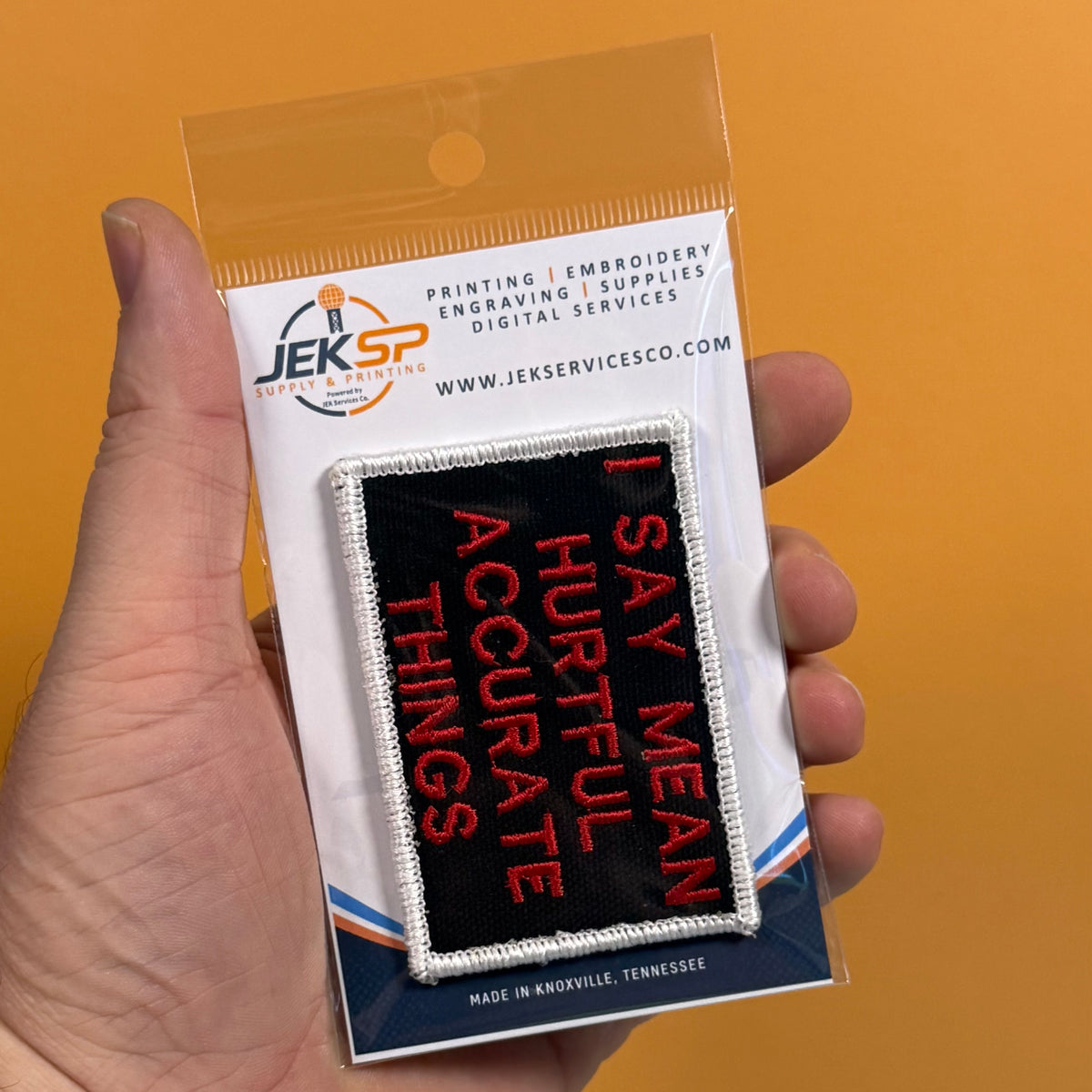 A hand holding a packaged embroidered patch that reads 'I SAY MEAN HURTFUL ACCURATE THINGS' in red on a black background, with a white stitched border. The package card shows the Jeksp Supplies brand and website, against a solid orange background.