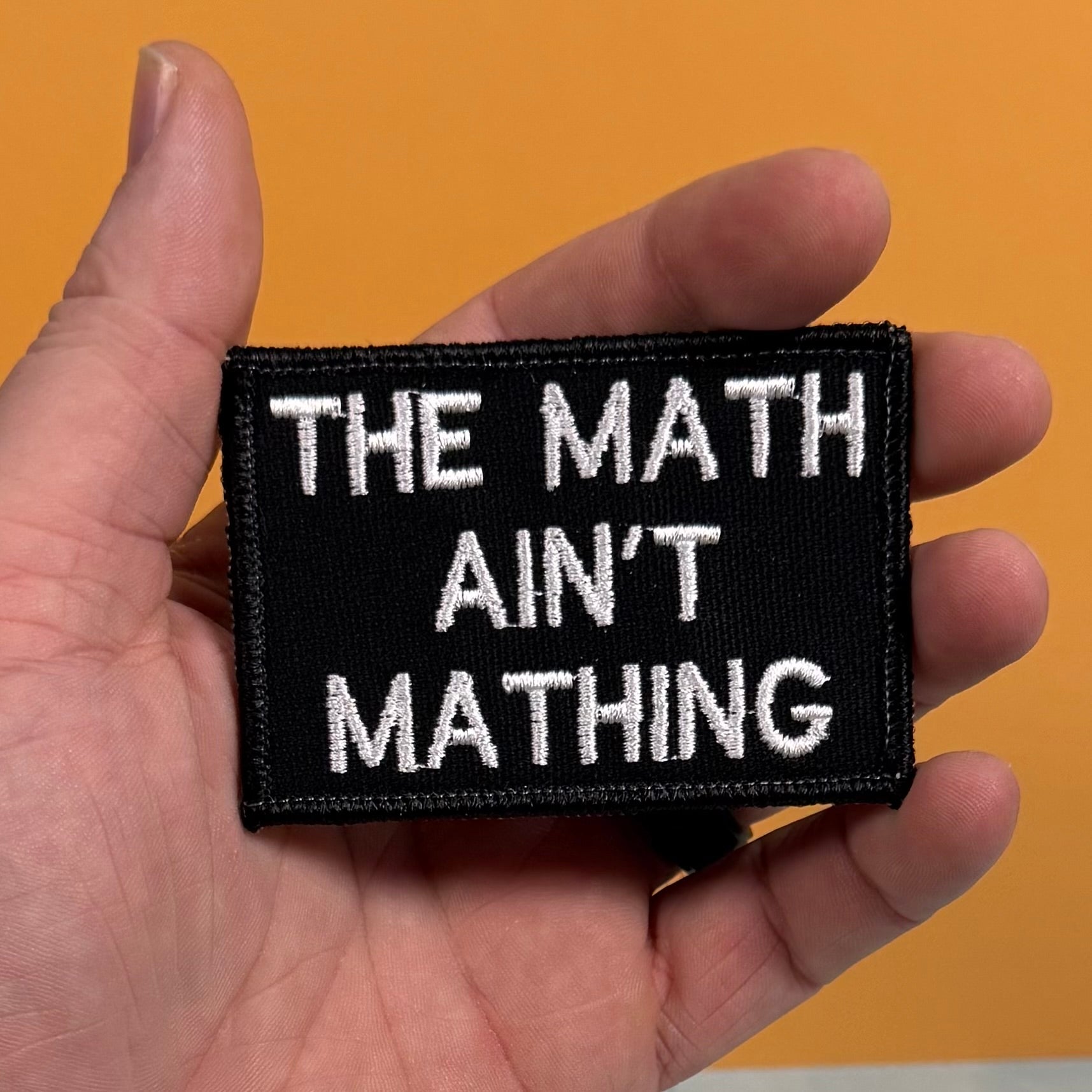 A hand holding a rectangular black embroidered patch with white text that reads 'THE MATH AIN'T MATHING' against a solid orange background.
