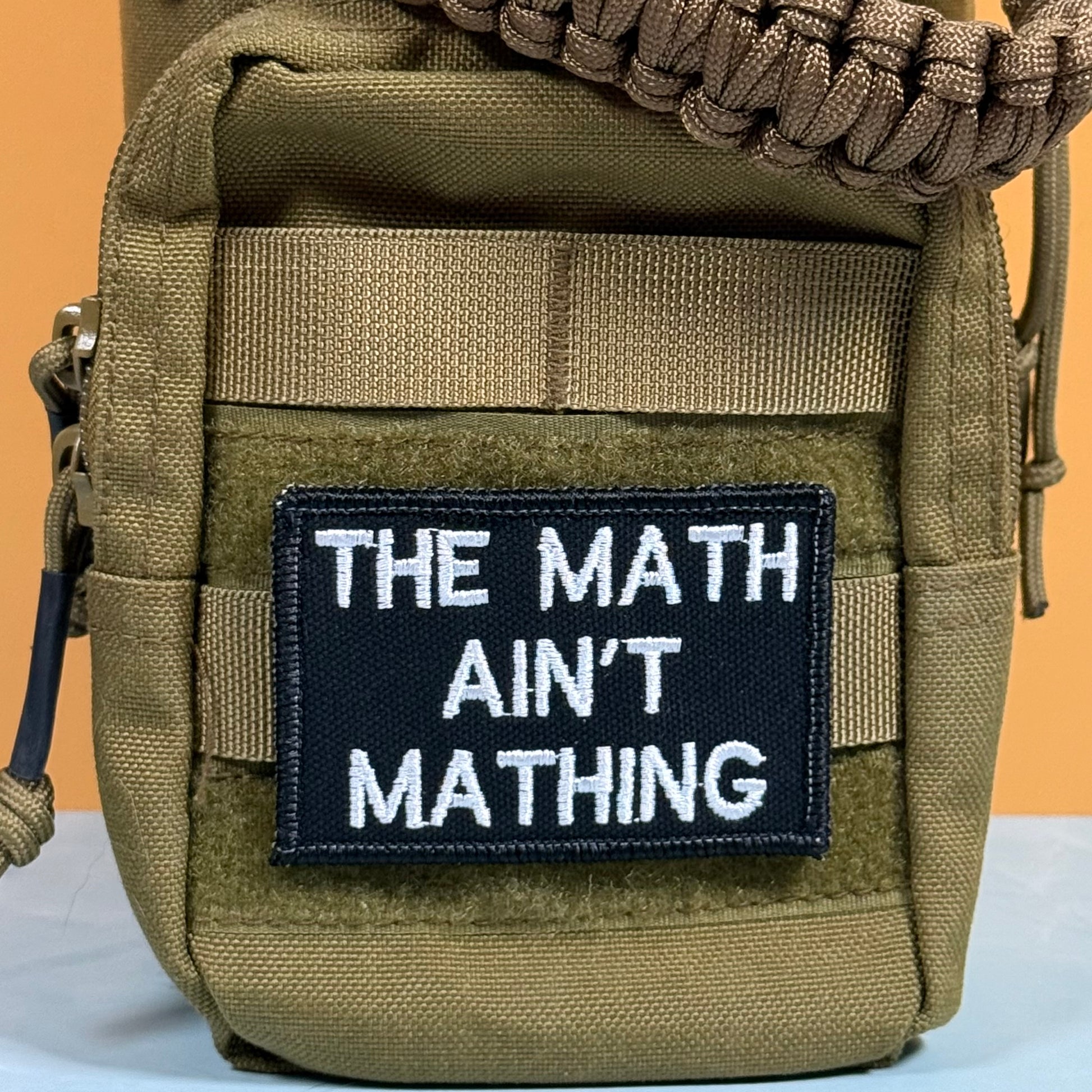 A close-up of a tactical utility pouch in olive drab/camo fabric, featuring a black rectangular patch with white text that reads 'THE MATH AIN'T MATHING'. The pouch has a tan woven strap across the top and side zippers, set against a split orange and white background.