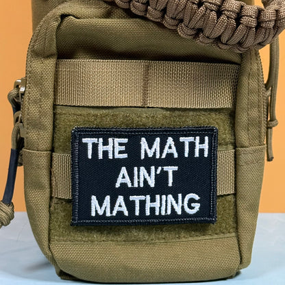 A close-up of a tactical utility pouch in olive drab/camo fabric, featuring a black rectangular patch with white text that reads 'THE MATH AIN'T MATHING'. The pouch has a tan woven strap across the top and side zippers, set against a split orange and white background.