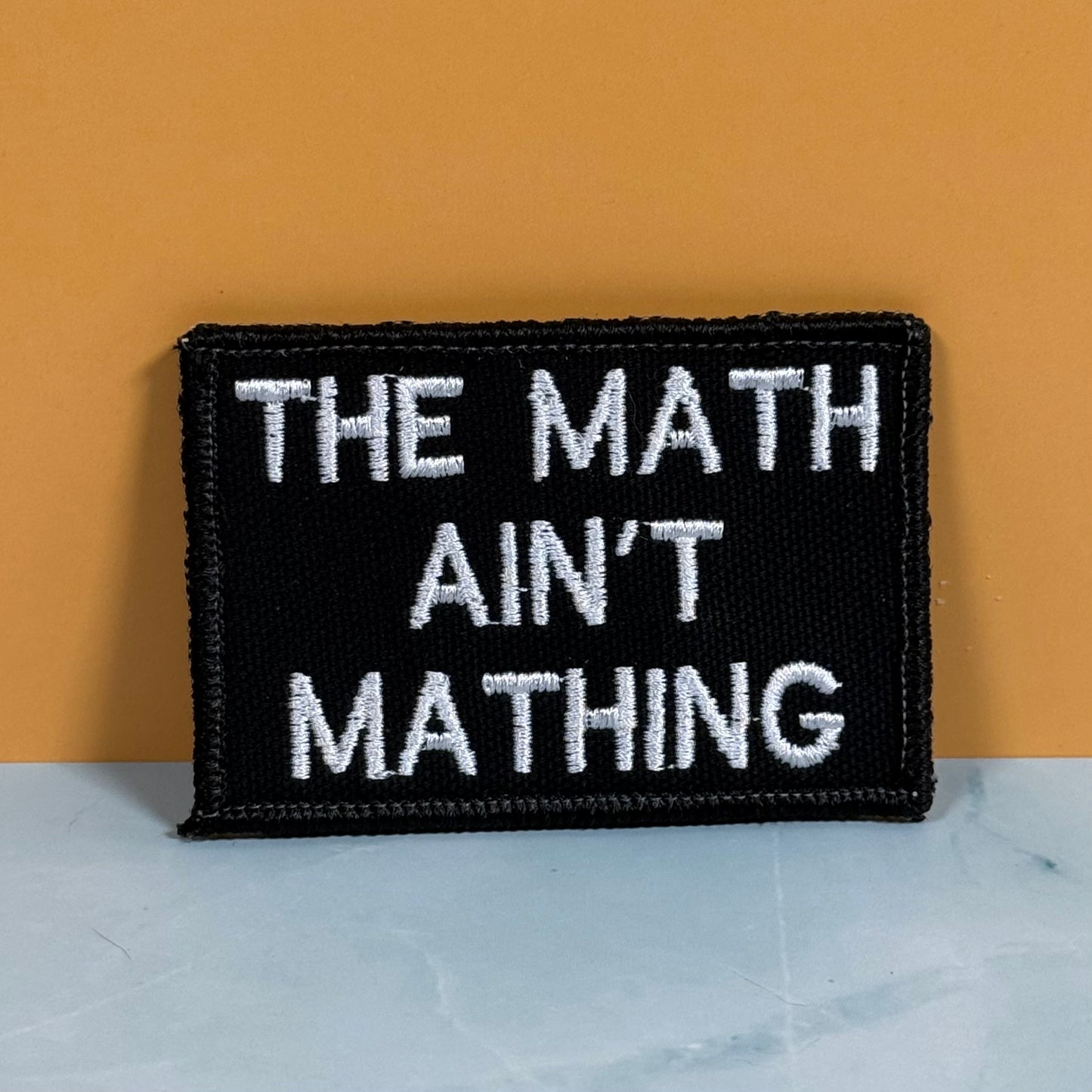 A rectangular black embroidered patch with white text reading 'THE MATH AIN'T MATHING' sits on a light marble-like surface against a warm orange background.