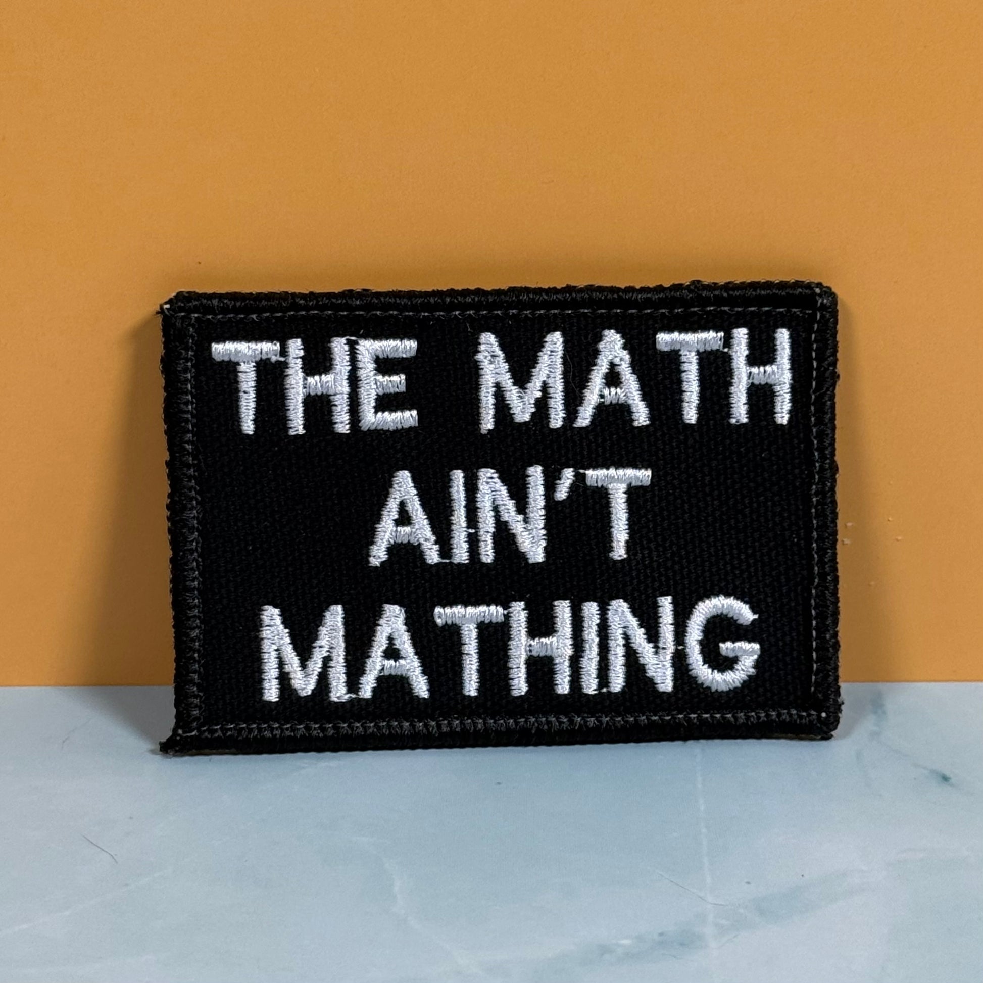 A rectangular black embroidered patch with white text reading 'THE MATH AIN'T MATHING' sits on a light marble-like surface against a warm orange background.