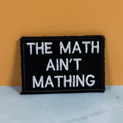 A rectangular black embroidered patch with white text reading 'THE MATH AIN'T MATHING' sits on a light marble-like surface against a warm orange background.