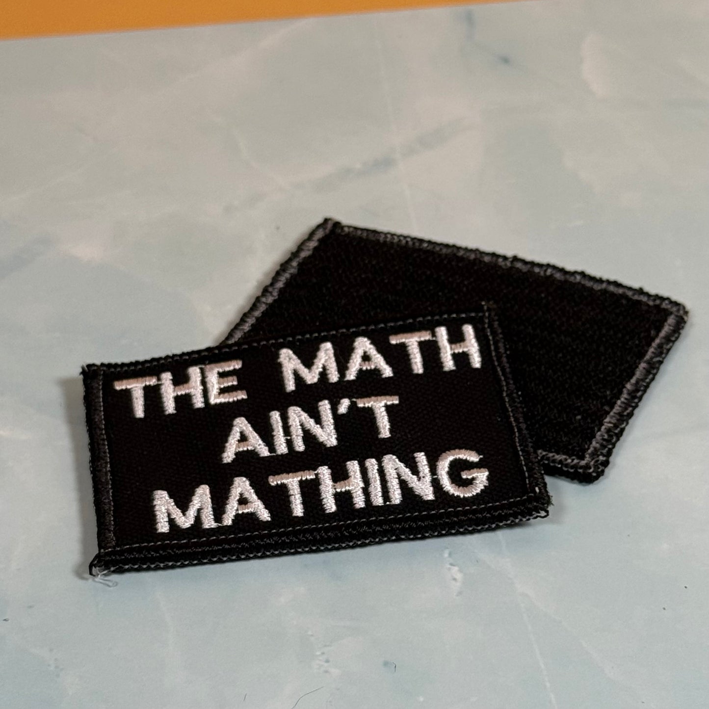Two black embroidered patches with white text reading 'THE MATH AIN'T MATHING' on a light gray marble-like surface.