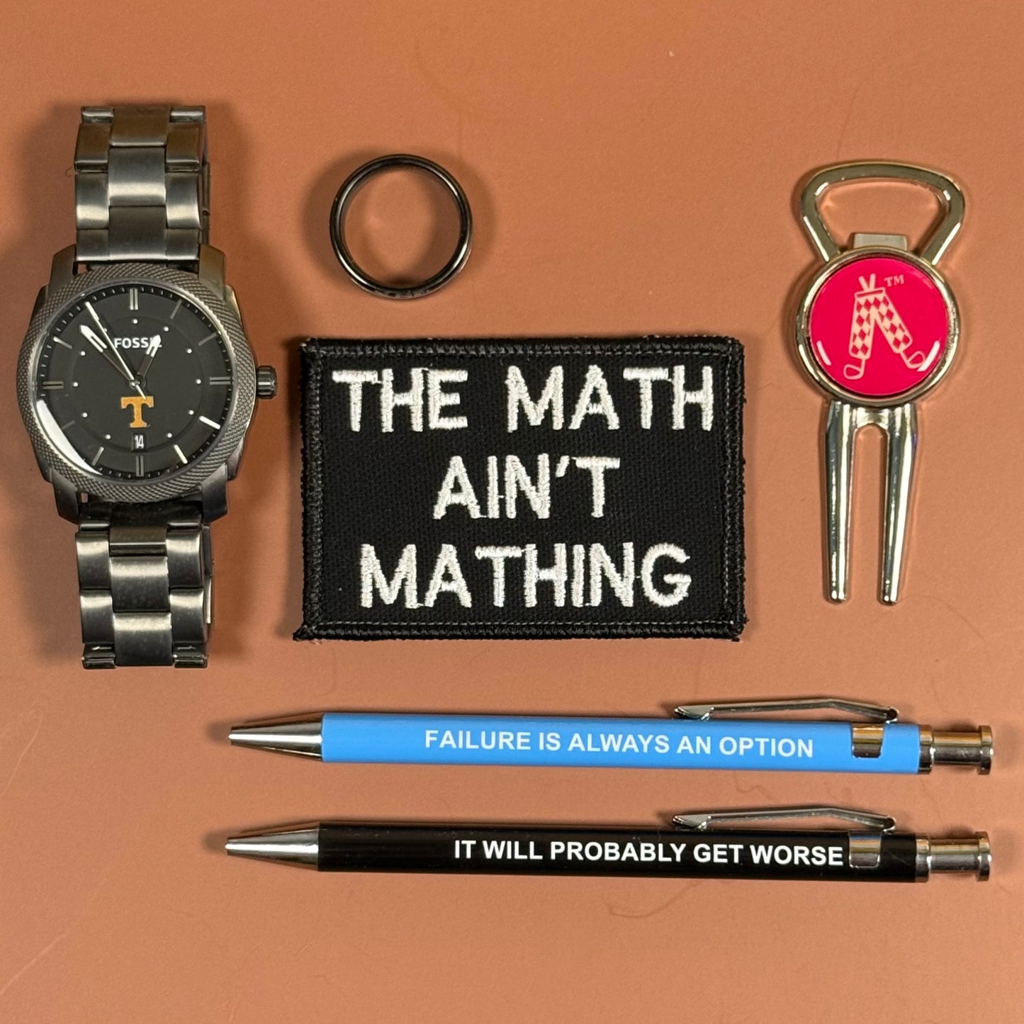 A flat-lay collection of collegiate-themed accessories on a solid terracotta background, including a silver watch, a black embroidered patch reading 'THE MATH AIN'T MATHING', a black ring, a silver keychain with a red Alabama logo, and two pens, one blue and one black, both printed with motivational text.