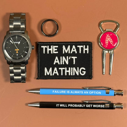 A flat-lay collection of collegiate-themed accessories on a solid terracotta background, including a silver watch, a black embroidered patch reading 'THE MATH AIN'T MATHING', a black ring, a silver keychain with a red Alabama logo, and two pens, one blue and one black, both printed with motivational text.