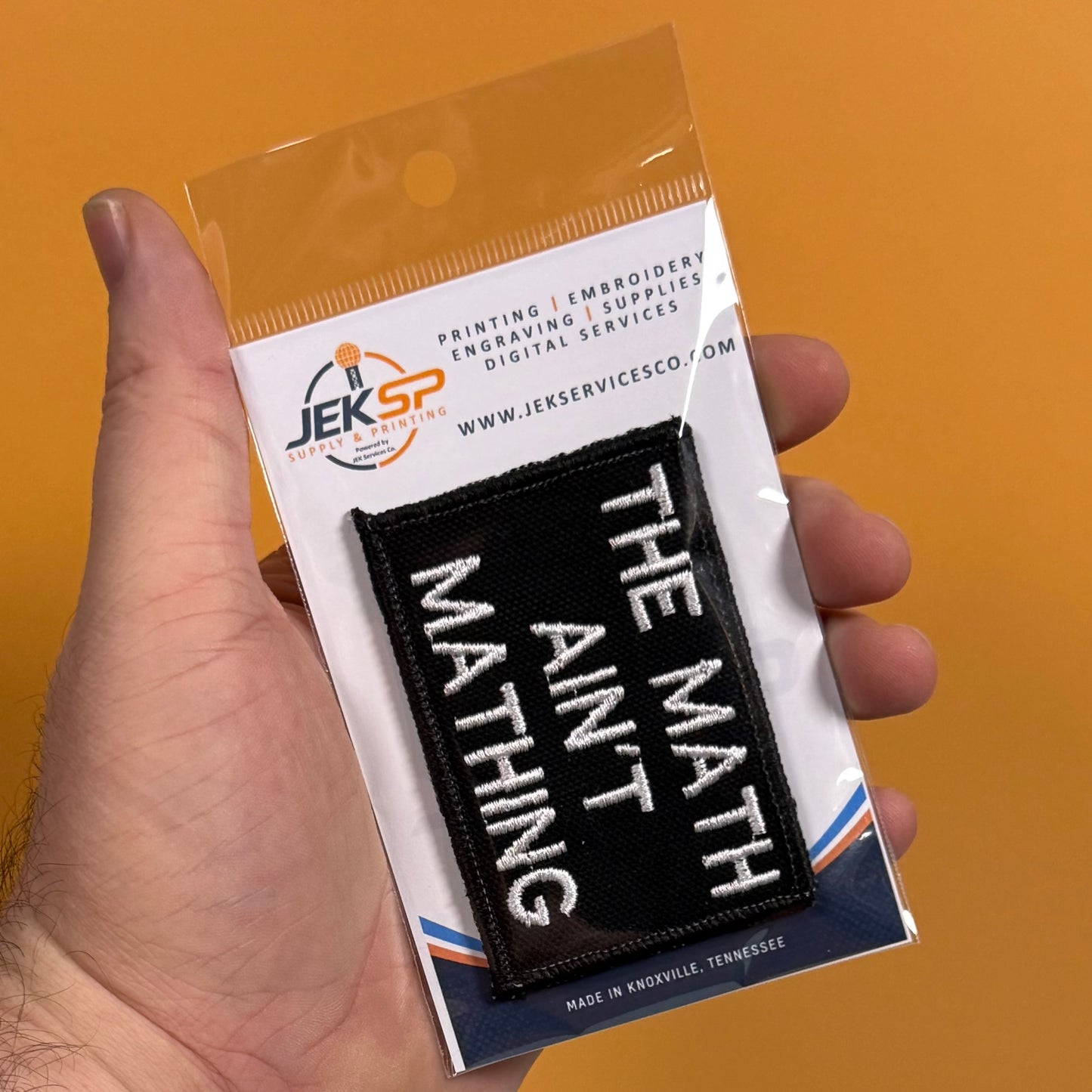 A hand holding a packaged black embroidered patch with white text reading 'THE MATH DOESN'T WANT ME' against an orange background. The backing card shows the Jeksp Service logo and product details.