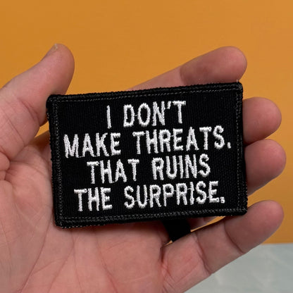 A hand holding a rectangular black embroidered patch with white text that reads, 'I DON'T MAKE THREATS. THAT RUINS THE SURPRISE.