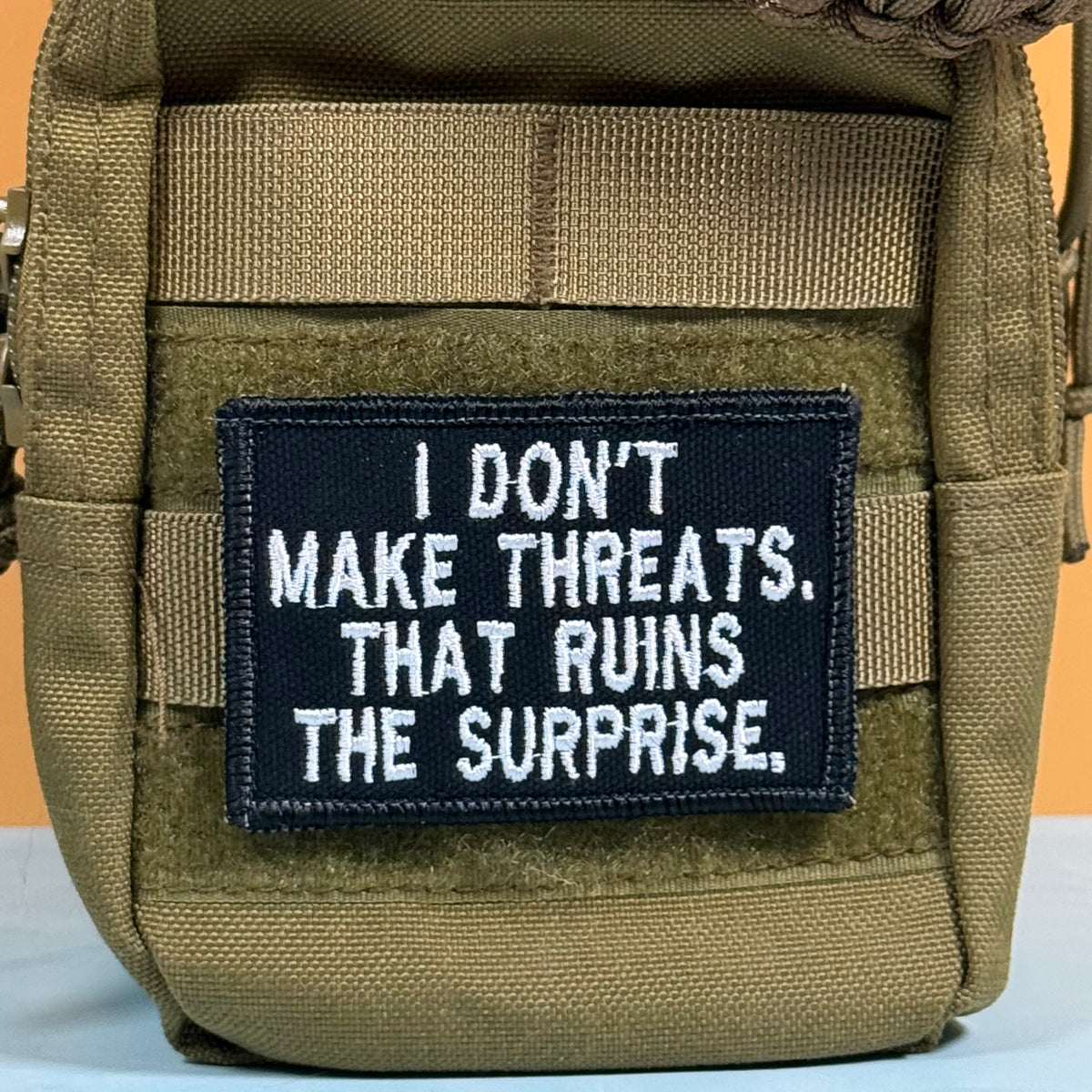 A close-up of a tactical pouch in olive drab green webbed material, featuring a rectangular black patch with white embroidered text that reads, 'I DON'T MAKE THREATS. THAT RUINS THE SURPRISE.