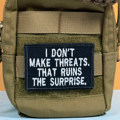 A close-up of a tactical pouch in olive drab green webbed material, featuring a rectangular black patch with white embroidered text that reads, 'I DON'T MAKE THREATS. THAT RUINS THE SURPRISE.