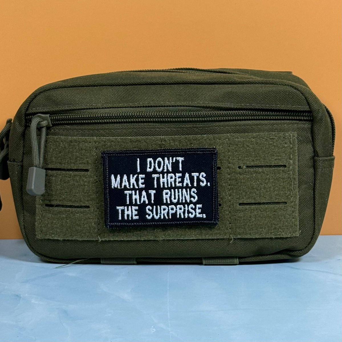 An olive green tactical pouch with a black rectangular patch that reads 'I DON'T MAKE THREATS. THAT RUINS THE SURPRISE.
