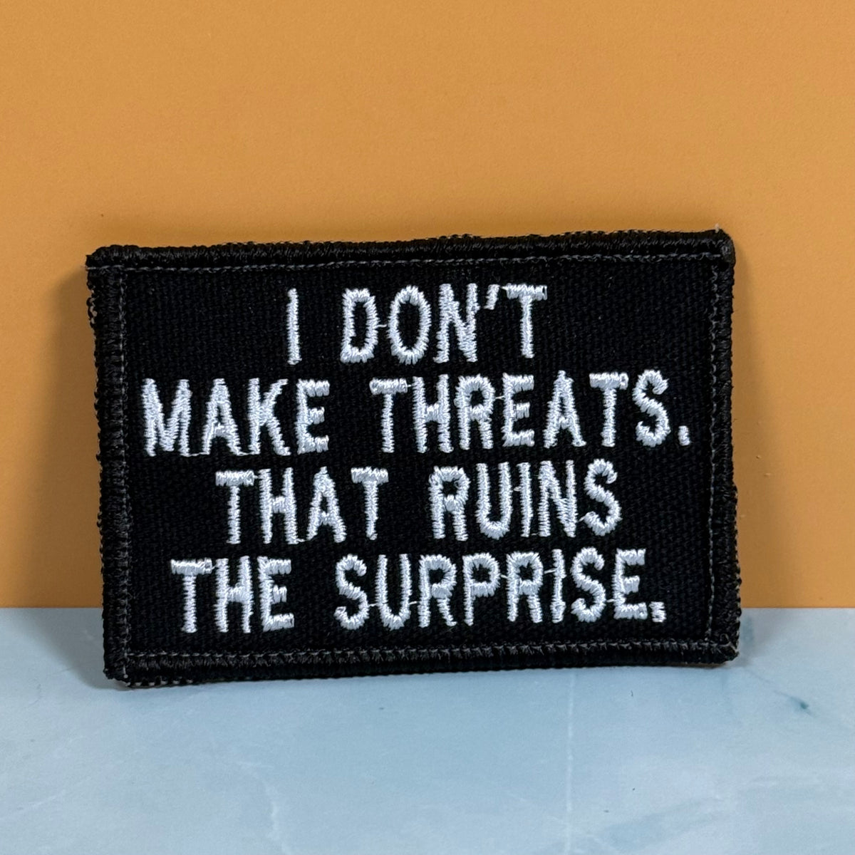 A rectangular black embroidered patch with white text reading 'I DON'T MAKE THREATS. THAT RUINS THE SURPRISE.