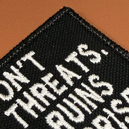 A close-up of a black embroidered patch with white text reading 'DON'T THREATEN THE EATS' on a warm brown background.