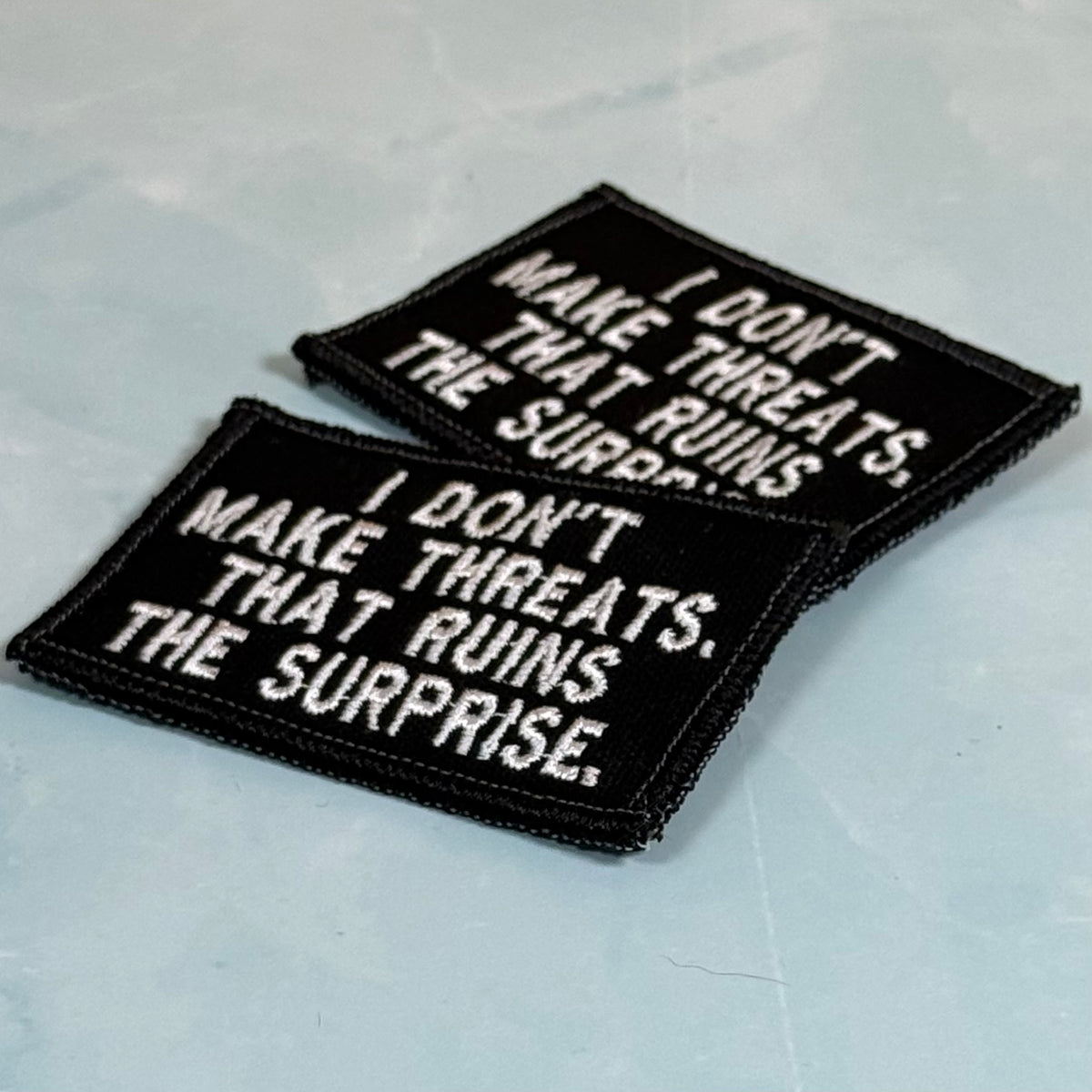 Two rectangular black embroidered patches with white text reading 'I DON'T MAKE THREATS. THAT RUINS THE SURPRISE.