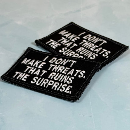 Two rectangular black embroidered patches with white text reading 'I DON'T MAKE THREATS. THAT RUINS THE SURPRISE.