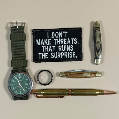 A flat-lay collection of vintage military surplus items on a light beige background, including a green nylon watch strap, a pocket knife, a bullet pen, a circular black ring, and a black embroidered patch with white text reading 'I DON'T MAKE THREATS. THAT RUINS THE SURPRISE.