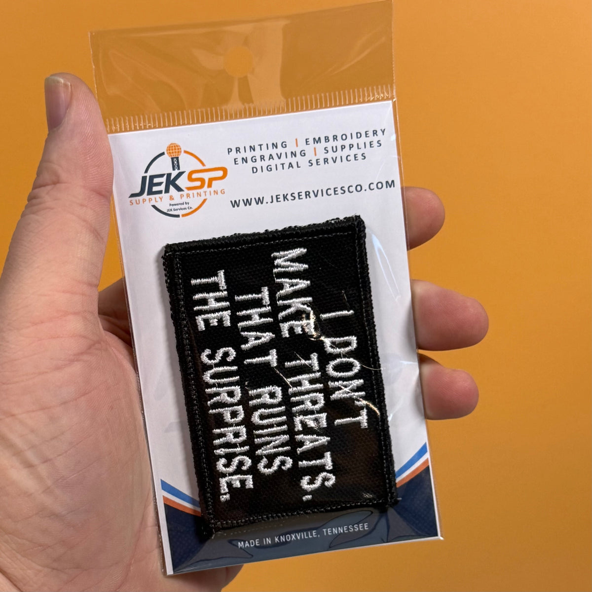 A hand holds a packaged black embroidered patch with white text reading, 'I DON'T MAKE THREATS THAT RUNS THE SURPRISE.' The backing card shows the brand JEKSP Supplies & Printing and includes website text, against a solid orange background.