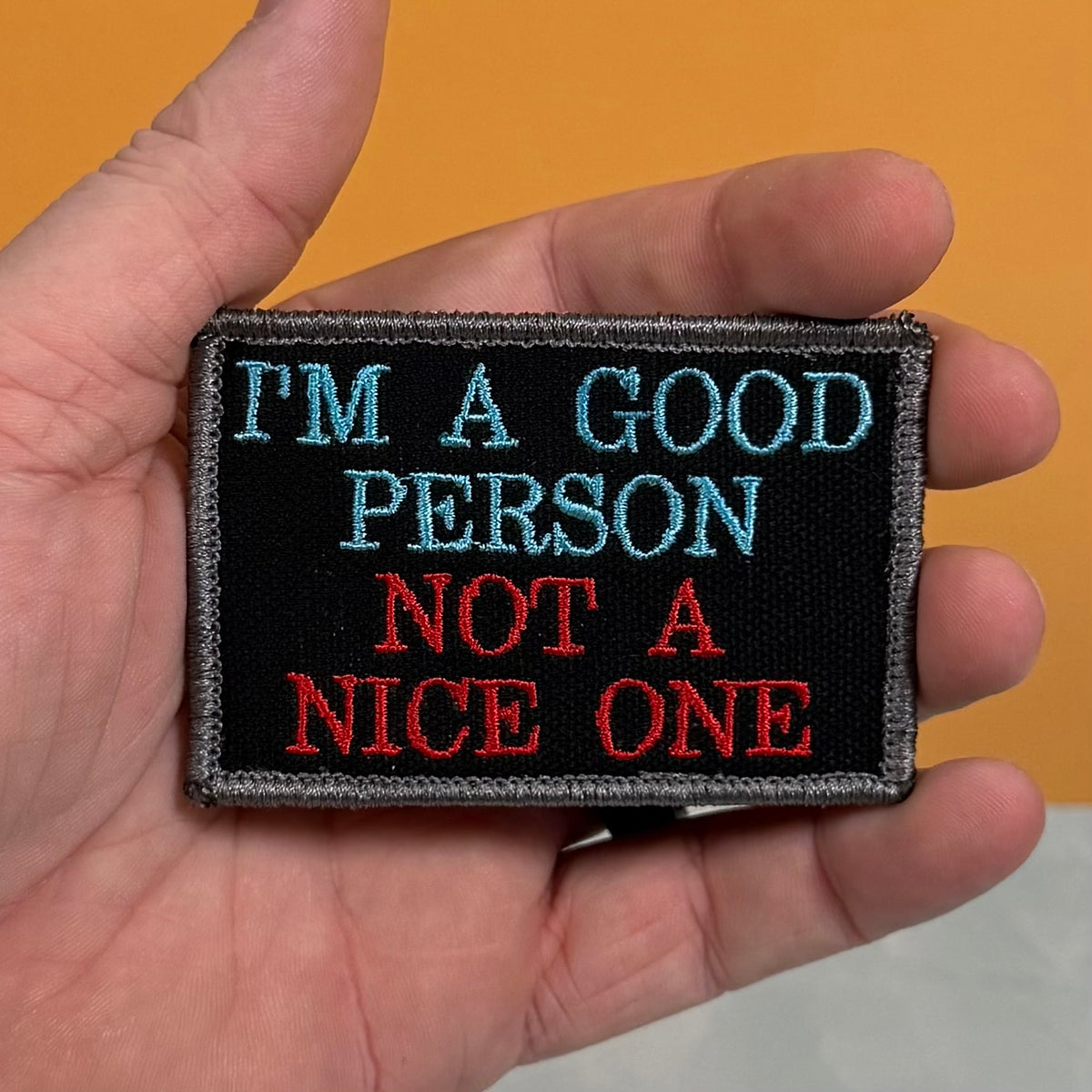 A hand holding a rectangular embroidered patch with the text 'I'M A GOOD PERSON NOT A NICE ONE' in light blue and red on a black background, set against an orange wall.