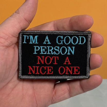 A hand holding a rectangular embroidered patch with the text 'I'M A GOOD PERSON NOT A NICE ONE' in light blue and red on a black background, set against an orange wall.