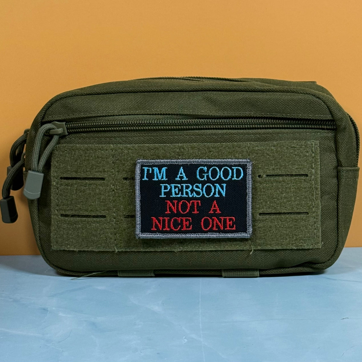 An olive green tactical pouch with a front zipper and a rectangular embroidered patch that reads 'I'M A GOOD PERSON NOT A NICE ONE' in blue and red text on a black background, set against a solid orange wall and light blue surface.