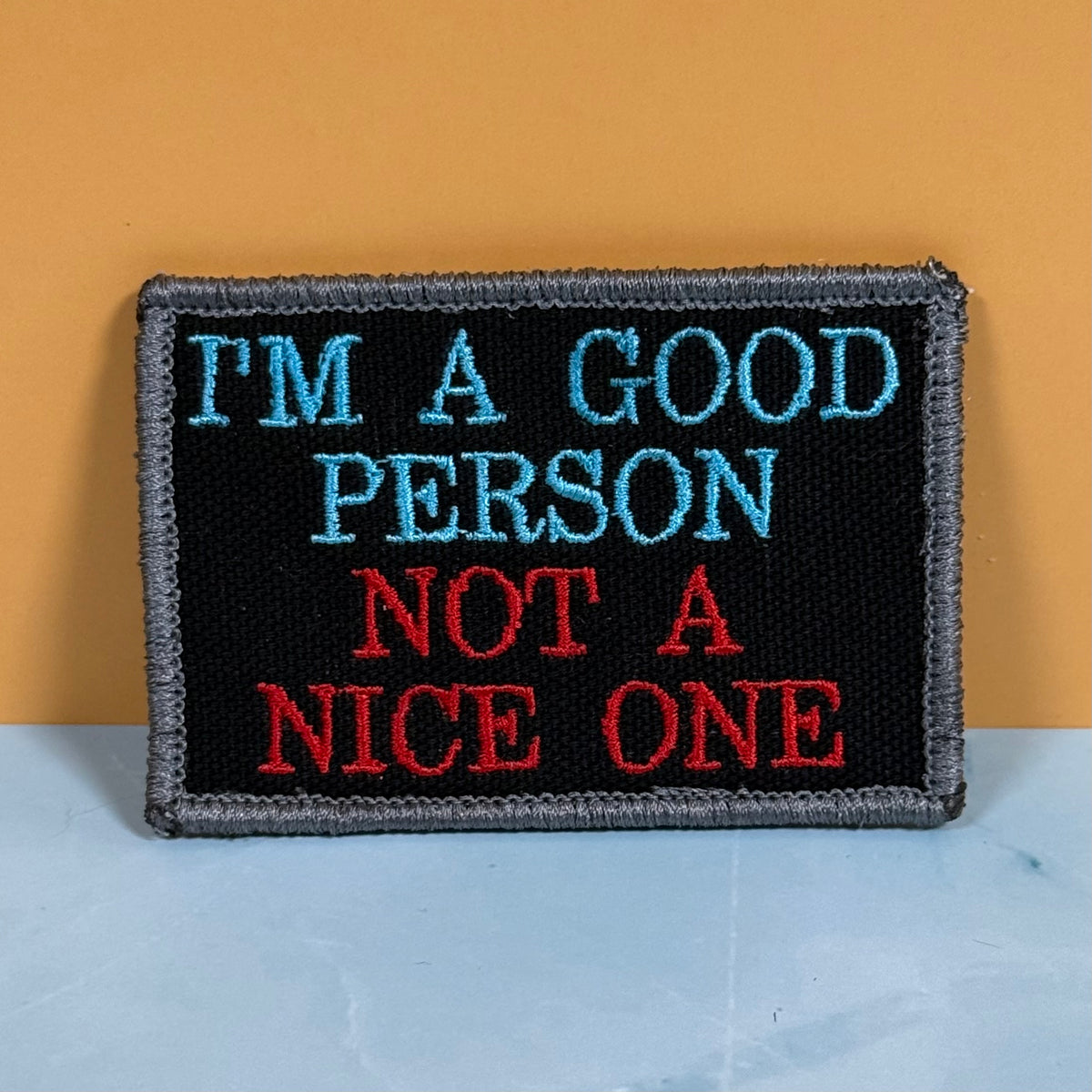 An embroidered rectangular patch with a black background and gray border, featuring the text 'I'M A GOOD PERSON NOT A NICE ONE' in light blue and red lettering, set against a two-tone orange and light blue background.