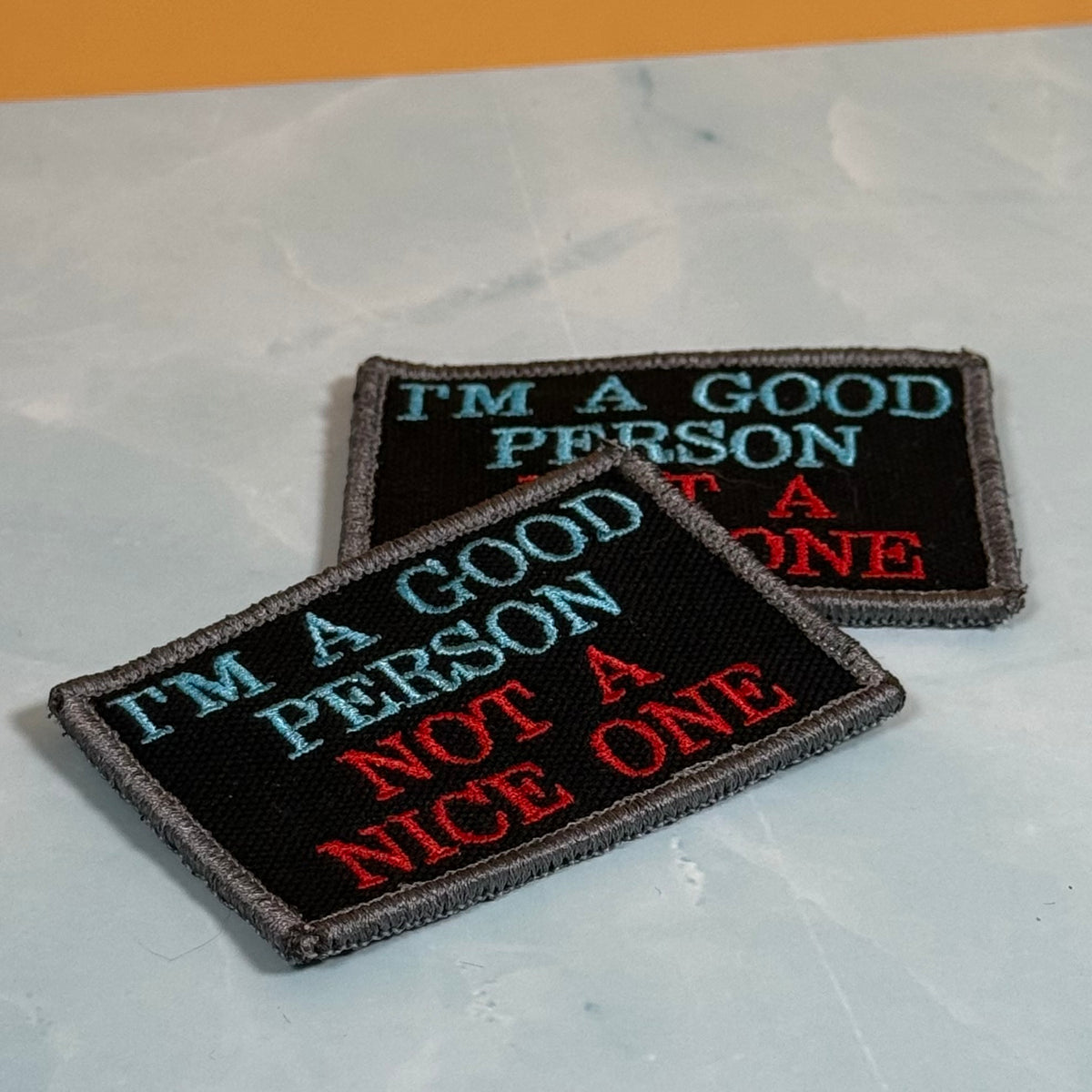 Two rectangular embroidered patches with black backgrounds and gray stitched borders, featuring the text 'I'M A GOOD PERSON NOT A ONE' in light blue and red lettering, displayed on a light gray marble-like surface.
