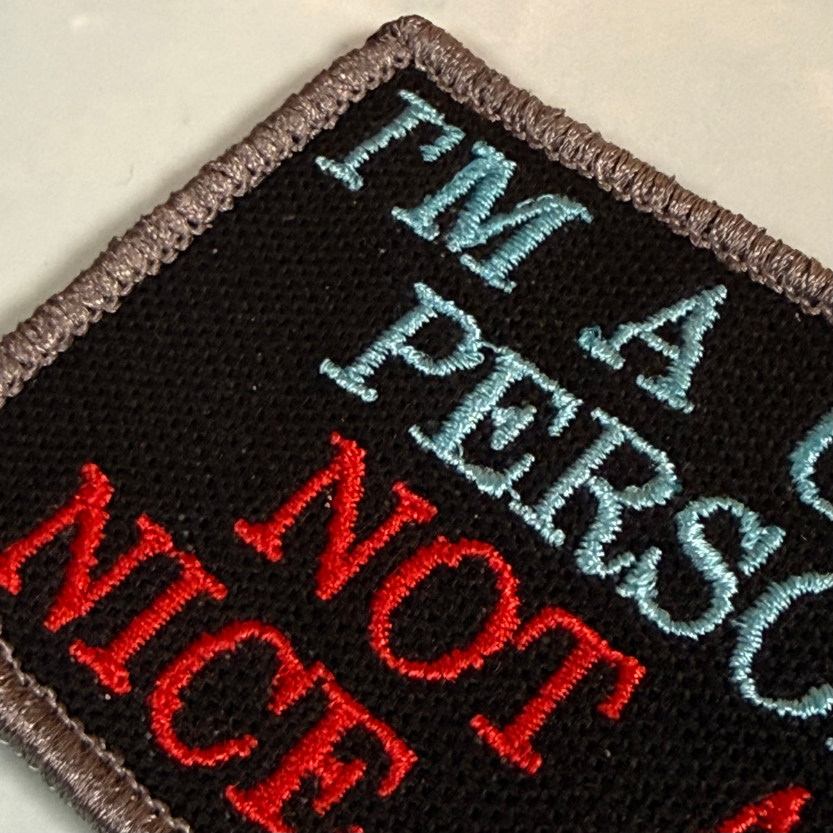 A close-up of a rectangular embroidered patch with a black background and brown stitched border. The patch features the text 'I'M A PERSON NOT NICE' in light blue and red embroidery.