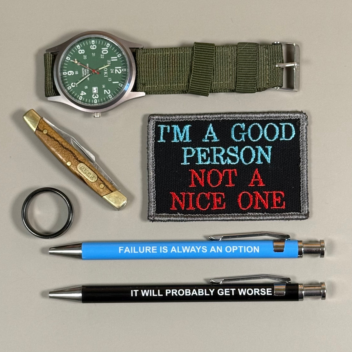 A flat-lay collection of novelty items on a light gray background, including a green military-style wristwatch, a folding pocket knife, a black patch with the text 'I'M A GOOD PERSON NOT A NICE ONE' in blue and red, a black keychain, a blue pen with the text 'FAILURE IS ALWAYS AN OPTION', and a black pen with the text 'IT WILL PROBABLY GET WORSE'.